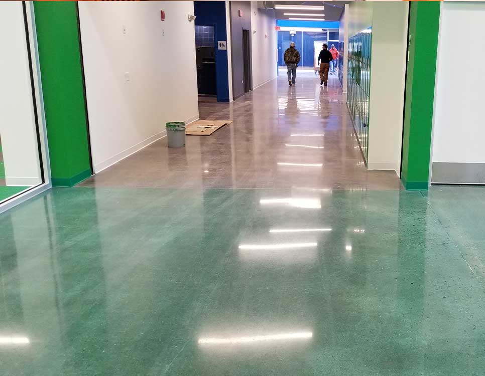 Dyed Concrete Flooring in a school hallway featuring a polished green concrete surface transitioning into natural gray, with lockers and students walking.